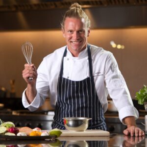 A 40-year-old American male chef with blond hair, wearing a white chef's jacket and apron, standing in a bright, modern kitchen with fresh ingredients on the counter.